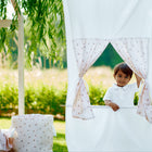 Ein kleines Kind lugt hinter dem KraftKids Puppentheater Kasperletheater Musselin Kirschen mit Kirschenmuster und Teleskopstange hervor, das draußen in einem sonnigen Garten umgeben von Grün und Bäumen aufgestellt ist.