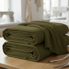 Zwei gefaltete KraftKids Wandpaneel abgerundet Doppelkrepp in olivgrün liegen gestapelt auf einem Holztisch in einem hellen Raum, im Hintergrund sind ein Maßband und unscharfes Nähmaterial zu sehen.