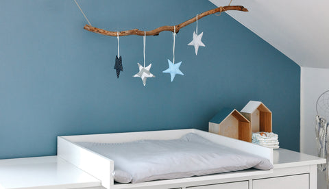 Eine minimalistische Wickelstation mit einem KraftKids Wickelaufsatz weiß für eine 160 cm HEMNES Kommode, gepolsterter Unterlage, Holzhäusern, gestapelten Windeln und einem handgefertigten Sternenmobile an einer beruhigenden blauen Wand.