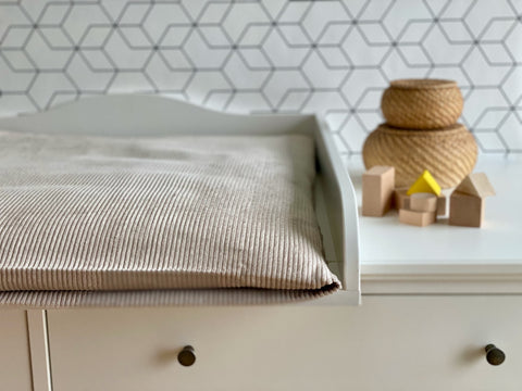 Ein weißer Wickeltisch mit einer KraftKids Bezug für Wickeltischauflage Cord Breitcord in Beige Taupe (100% Baumwolle), gepaart mit Holzblöcken und zwei geflochtenen Körben, vor einer sechseckigen Wand.