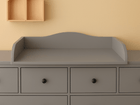Eine graue Kommode mit sechs Schubladen und schwarzen Knöpfen wird von dem Wickelaufsatz grau passend für 160 cm breite HEMNES Kommode mit Blende gekrönt. Darüber ist an der beigen Wand ein KraftKids-Holzregal in Form von zwei Hausdächern angebracht.