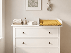 Eine weiße HEMNES Kommode mit einem KraftKids Wickelaufsatz (weiß, passend für 108 cm breite Kommode, Wickelfläche 50×70 cm) fasst Handtücher und eine gelbe Wickelauflage. Oben schmücken KraftKids Blattkunst und ein Tieraufhänger das sanft beleuchtete Zimmer.
