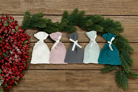 Fünf handgemachte KraftKids Musselin-Stoffsäckchen in Grün, Grau, Rosa und Weiß mit goldenen Punkten und weißen Bändern liegen arrangiert auf Holz mit Tannenzweigen - perfekt für deinen Adventskalender.