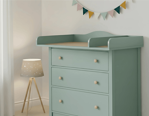 In einem sanft beleuchteten Zimmer steht eine mintgrüne Kommode mit vier Schubladen, auf der der KraftKids Wickelaufsatz XXL pastellgrün extra breit 108 cm mit Trennfach für HEMNES steht. Eine dreibeinige Lampe mit Sternenmuster und eine pastellfarbene Wimpelkette setzen charmante Akzente.