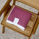 Eine KraftKids Reisewickelunterlage Leinen magenta dunkel liegt zusammengefaltet auf einem Holzstuhl mit geflochtenem Rohrsitz, der auf einem hellen Teppich steht.