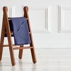 Eine kleine hölzerne Stufenleiter mit einer gefalteten KraftKids Babydecke für Babyschale Sommer Leinen blau dunkelblau, die über eine Stufe drapiert ist, steht auf einem hellen Holzboden vor einer weißen Wand mit Zierleisten.