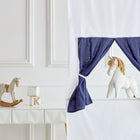 Ein weiß-blaues Spielzelt mit einem Spielzeug-Einhorn steht neben einem weißen Tisch mit einem Holzschaukelpferd, Buchstabenblöcken und dem KraftKids Puppentheater Kasperletheater Leinen grau Pusteblumen inkl. Teleskopstange. Teleskopstange, vor einer weiß getäfelten Wand.