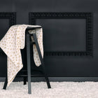 Die KraftKids Babydecke Musselin Zweige braun ist über einen schwarzen Holzhocker drapiert und steht vor einer dunklen Wand mit Leisten und einem weißen, flauschigen Teppich darunter.