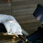 Eine Person mit schwarzen Ärmeln schiebt einen Kinderwagen im Freien auf einer Holzfläche, an dessen Griff der KraftKids Kinderwagenmuff Musselin Kirschen befestigt ist.