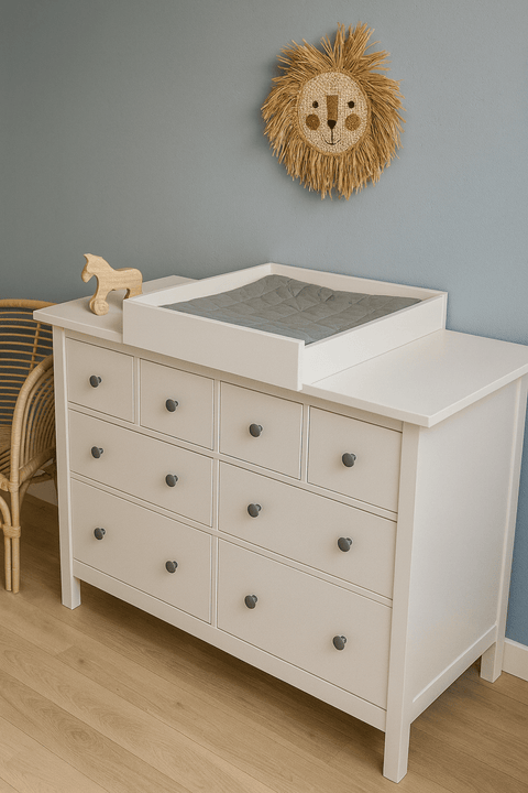 Eine weiße Kommode mit neun Schubladen und dem KraftKids Wickelaufsatz weiß zum Quer-Wickeln für HEMNES steht neben einem Holzspielzeugpferd. Darüber hängt eine Wanddekoration mit einem Löwen aus Rattan, und ein Korbstuhl lugt von der Seite herein.