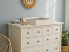 In einem hellen, sonnendurchfluteten Raum sorgen ein KraftKids Wickelaufsatz weiß zum Quer-Wickeln auf einer HEMNES Kommode mit neun Schubladen, ordentlich gefaltete Kleidung, ein Wanddekor mit Löwengesicht, ein Rattanstuhl und eine Topfpflanze für platzsparenden Stil.