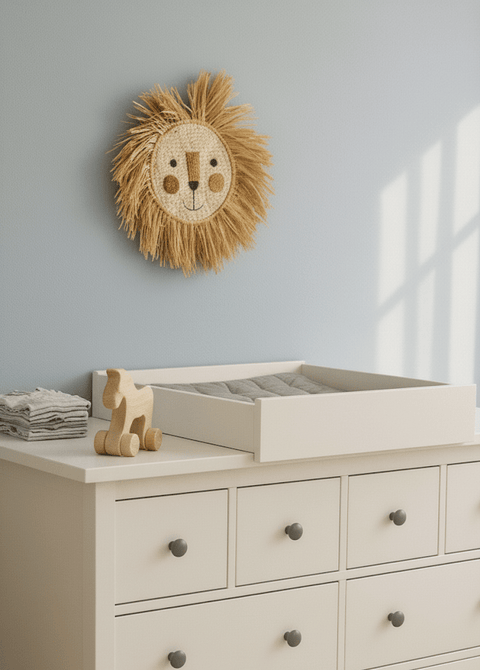 In einem Kinderzimmer stehen eine weiße Kommode mit dem KraftKids Wickelaufsatz weiß zum Quer-Wickeln für breite und schmale HEMNES Kommode, ordentlich gefaltete Handtücher, ein Holzspielzeugpferd und ein dekorativer Wandbehang mit Löwengesicht an der hellblauen Wand.