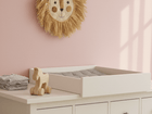 Ein Kinderzimmer mit einer rosafarbenen Wand, einem Wandbehang mit Löwengesicht und einem KraftKids Wickelaufsatz weiß zum Quer-Wickeln auf einer weißen HEMNES Kommode mit dunklen Knöpfen. Ein Holzspielzeugpferd und gefaltete Tücher liegen daneben. Sonnenlicht wirft Rechteckschatten an die Wand.