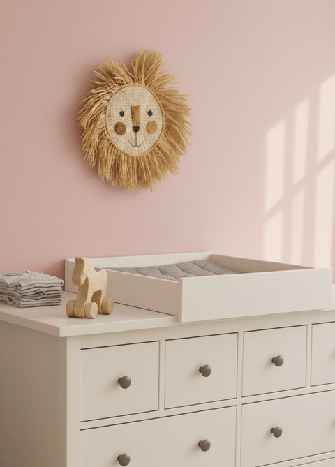 Ein Kinderzimmer mit einer rosafarbenen Wand, einem Wandbehang mit Löwengesicht und einem KraftKids Wickelaufsatz weiß zum Quer-Wickeln auf einer weißen HEMNES Kommode mit dunklen Knöpfen. Ein Holzspielzeugpferd und gefaltete Tücher liegen daneben. Sonnenlicht wirft Rechteckschatten an die Wand.