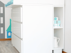 In einem hellen, minimalistischen Zimmer mit Holzfußboden steht das KraftKids Stauraumregal für Wickeltisch in weiß, passend zur MALM Kommode mit 3 oder 4 Schubladen, in der gefaltete Decken, ein Krug und Deko-Boxen Platz finden.