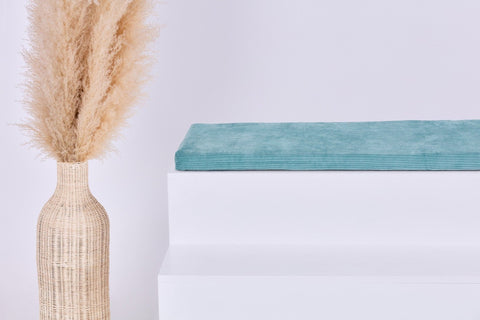 Eine geflochtene Vase mit Pampasgras steht neben einer KraftKids GmbH Sitzauflage für Ikea TROFAST Cord Breitcord Mint, gekrönt von einem gefalteten tealfarbenen Handtuch, vor einem schlichten hellen Hintergrund.