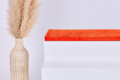 Eine hohe geflochtene Vase mit beigem Pampasgras steht neben einem weißen Stufenpodest, auf dem die KraftKids GmbH Sitzauflage für Ikea STUVA/SMÅSTAD Cord Breitcord Orange Reinorange steht. Der Hintergrund ist schlicht und minimalistisch.