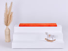 Ein weißes dreistufiges Podest mit einer KraftKids GmbH Sitzauflage für Ikea TROFAST Cord Breitcord Orange Reinorange auf der obersten Stufe steht zwischen einer beigen geflochtenen Vase mit Pampasgras (links) und einem kleinen weißen Holzschaukelpferd (rechts).