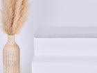 Eine hohe Korbvase mit beigem Pampasgras steht neben einer weißen, minimalistischen Bank, auf der die KraftKids GmbH Sitzauflage für Ikea TROFAST Musselin grau Pusteblumen steht. Der Hintergrund ist einfarbig weiß.