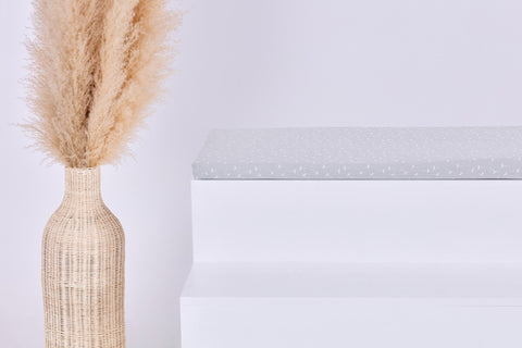 Eine hohe geflochtene Vase mit Pampasgras steht neben einem weißen, gestuften IKEA STUVA/SMÅSTAD Regal mit der KraftKids GmbH Sitzauflage für Ikea STUVA/SMÅSTAD Musselin grau Pusteblumen. Der Hintergrund ist schlicht und minimalistisch.