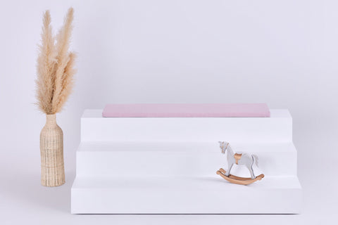 Eine minimalistische Szene zeigt ein weißes dreistufiges Podest mit einem KraftKids GmbH Sitzauflage für Ikea TROFAST Doppelkrepp Rosa Kissen, ein hölzernes Schaukelpferd davor und eine hohe geflochtene Vase mit Pampasgras auf der linken Seite vor einem hellen Hintergrund.