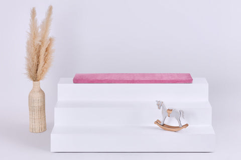 Eine dreistufige weiße IKEA TROFAST Sitzbank mit einem KraftKids GmbH Sitzauflage für Ikea TROFAST Cord Breitcord Rosa Kissen auf der obersten Stufe, einer Vase mit Pampasgras auf der linken Seite und einem kleinen weißen Schaukelpferd auf der unteren rechten Seite.