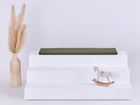 Auf einem weißen, dreistufigen Podest steht die KraftKids GmbH Sitzauflage für Ikea TROFAST Doppelkrepp in Herbstgrün. Links steht eine geflochtene Vase mit Pampasgras, rechts sitzt ein kleines weißes und hölzernes Schaukelpferd vor einem hellen Hintergrund.
