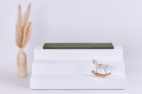 Auf einem weißen, dreistufigen Podest steht die KraftKids GmbH Sitzauflage für Ikea TROFAST Doppelkrepp in Herbstgrün. Links steht eine geflochtene Vase mit Pampasgras, rechts sitzt ein kleines weißes und hölzernes Schaukelpferd vor einem hellen Hintergrund.