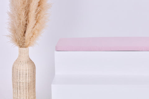 Eine hohe geflochtene Vase mit beigem Pampasgras steht neben einer modernen weißen Bank mit der KraftKids GmbH Sitzauflage für Ikea TROFAST Doppelkrepp Rosa vor einem schlichten weißen Hintergrund.