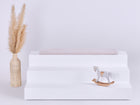Eine minimalistische Szene mit einem dreistufigen weißen Podest, auf dem die KraftKids GmbH Sitzauflage für Ikea TROFAST Cord Breitcord in Beige Taupe steht. Ein kleines hölzernes Schaukelpferd steht auf der untersten Ebene, während links eine hohe Vase mit getrocknetem Pampasgras steht.