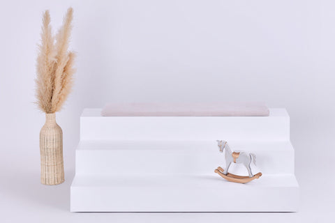 Eine minimalistische Szene mit einem dreistufigen weißen Podest, auf dem die KraftKids GmbH Sitzauflage für Ikea TROFAST Cord Breitcord in Beige Taupe steht. Ein kleines hölzernes Schaukelpferd steht auf der untersten Ebene, während links eine hohe Vase mit getrocknetem Pampasgras steht.
