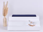 Ein weißes dreistufiges Podest mit einem KraftKids GmbH Sitzauflage für Ikea TROFAST Musselin dunkelblau Kissen auf der Oberseite, einem Schaukelpferd auf der untersten Stufe und einer hohen geflochtenen Vase mit Pampasgras auf der linken Seite, alles vor einem schlichten hellen Hintergrund.