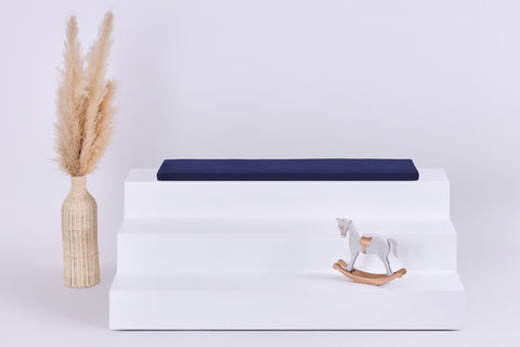 Ein weißes dreistufiges Podest mit einem KraftKids GmbH Sitzauflage für Ikea TROFAST Musselin dunkelblau Kissen auf der Oberseite, einem Schaukelpferd auf der untersten Stufe und einer hohen geflochtenen Vase mit Pampasgras auf der linken Seite, alles vor einem schlichten hellen Hintergrund.