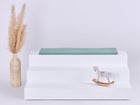 Eine geflochtene Vase mit Pampasgras, ein weißes dreistufiges Podest mit der KraftKids GmbH Sitzauflage für Ikea TROFAST Doppelkrepp Grün Jade und ein kleines Holzschaukelpferd vor hellem Hintergrund.