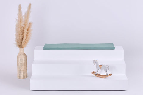 Eine geflochtene Vase mit Pampasgras, ein weißes dreistufiges Podest mit der KraftKids GmbH Sitzauflage für Ikea TROFAST Doppelkrepp Grün Jade und ein kleines Holzschaukelpferd vor hellem Hintergrund.