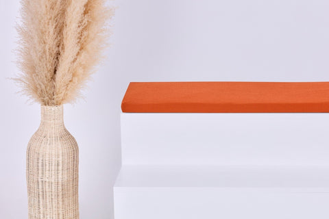 Eine hohe, geflochtene Vase mit beigem Pampasgras steht neben einer weißen Treppe mit der KraftKids GmbH Sitzauflage für Ikea KALLAX Regal in Doppelkrepp Rot Herbstrot auf der obersten Stufe, vor einem schlichten hellen Hintergrund.