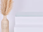 Eine hohe, geflochtene Vase mit beigem Pampasgras steht neben einer minimalistischen weißen Bank mit dem Kissen KraftKids GmbH Sitzauflage für Ikea TROFAST Musselin mint Pusteblumen. Der Hintergrund ist schlicht und hell gehalten.