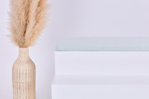 Eine geflochtene Vase mit hohem beigem Pampasgras steht neben einem weißen Stufenpodest, auf dem die KraftKids GmbH Sitzauflage für Ikea STUVA/SMÅSTAD Musselin mint Pusteblumen steht. Der schlichte Hintergrund unterstreicht die minimalistische Szene.
