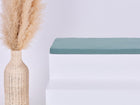Eine hohe geflochtene Vase mit Pampasgras steht neben einer minimalistischen weißen Bank mit einem KraftKids GmbH Sitzauflage für Ikea TROFAST Musselin nilblau Kissen vor einem schlichten hellen Hintergrund.