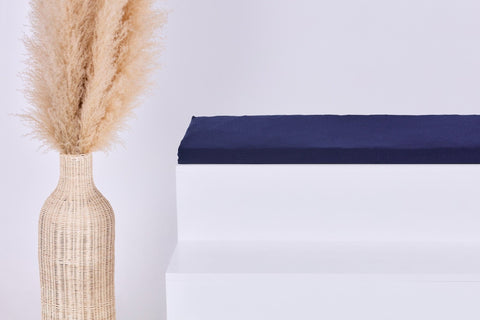 Eine hohe geflochtene Vase mit beigem Pampasgras steht neben einem weißen Stufenpodest, auf dem das KraftKids GmbH Sitzauflage für Ikea TROFAST Musselin dunkelblau Kissen liegt, vor einem schlichten hellen Hintergrund.