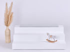 Auf einem weißen, dreistufigen Display steht die KraftKids GmbH Sitzauflage für Ikea TROFAST Musselin grau Pusteblumen neben einer beigen Vase mit Pampasgras und einem kleinen weißen Holzschaukelpferd. Der Hintergrund ist schlicht und hell.