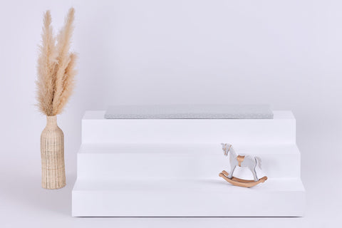 Auf einem weißen, dreistufigen Display steht die KraftKids GmbH Sitzauflage für Ikea TROFAST Musselin grau Pusteblumen neben einer beigen Vase mit Pampasgras und einem kleinen weißen Holzschaukelpferd. Der Hintergrund ist schlicht und hell.