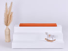 Ein weißes, dreistufiges Display mit einer roten KraftKids GmbH Sitzauflage für IKEA TROFAST Doppelkrepp Herbstrot auf der obersten Stufe, einer Vase mit beigem Pampasgras auf der linken Seite und einem kleinen Holzschaukelpferd auf der rechten Seite vor einem hellen Hintergrund.