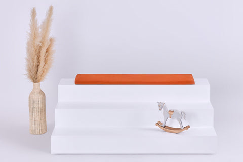 Ein weißes, dreistufiges Display mit einer roten KraftKids GmbH Sitzauflage für IKEA TROFAST Doppelkrepp Herbstrot auf der obersten Stufe, einer Vase mit beigem Pampasgras auf der linken Seite und einem kleinen Holzschaukelpferd auf der rechten Seite vor einem hellen Hintergrund.