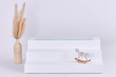 Ein weißer, dreistöckiger Ständer hält eine neuwertige Pusteblumen Sitzauflage für Ikea TROFAST von KraftKids GmbH, mit einer beigen Vase mit Pampasgras links und einem kleinen hölzernen Schaukelpferd unten rechts; der Hintergrund ist schlicht und hell.