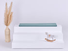 Ein weißer dreistöckiger Aufsteller mit einer KraftKids GmbH Sitzauflage für Ikea TROFAST Musselin nilblau oben, einer Korbvase mit getrocknetem Pampasgras links und einem kleinen weißen Holzschaukelpferd rechts, alles vor einem schlichten Hintergrund.