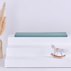 Ein weißer dreistöckiger Aufsteller mit einer KraftKids GmbH Sitzauflage für Ikea TROFAST Musselin nilblau oben, einer Korbvase mit getrocknetem Pampasgras links und einem kleinen weißen Holzschaukelpferd rechts, alles vor einem schlichten Hintergrund.