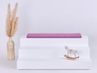 Eine minimalistische Szene mit einem dreistufigen weißen Aufsteller, auf dem die KraftKids GmbH Sitzauflage für Ikea TROFAST Musselin purpur steht, einer geflochtenen Vase mit getrocknetem Pampasgras auf der linken Seite und einem kleinen Holzschaukelpferd auf der rechten Seite.