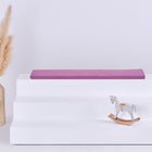 Eine minimalistische Szene mit einem dreistufigen weißen Aufsteller, auf dem die KraftKids GmbH Sitzauflage für Ikea TROFAST Musselin purpur steht, einer geflochtenen Vase mit getrocknetem Pampasgras auf der linken Seite und einem kleinen Holzschaukelpferd auf der rechten Seite.