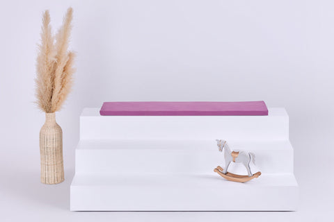 Eine minimalistische Szene mit einem dreistufigen weißen Aufsteller, auf dem die KraftKids GmbH Sitzauflage für Ikea TROFAST Musselin purpur steht, einer geflochtenen Vase mit getrocknetem Pampasgras auf der linken Seite und einem kleinen Holzschaukelpferd auf der rechten Seite.